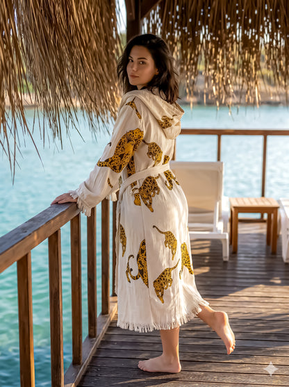 Woman in a white robe with gold animal prints standing on a wooden deck by a body of water.