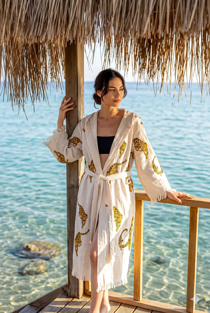 Woman in a white robe with gold patterns standing by a wooden deck with a thatched roof, overlooking clear blue water.