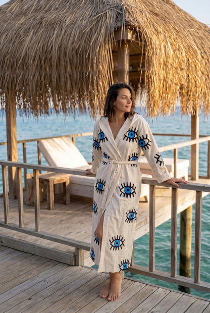 Woman in a robe with eye pattern standing on a wooden deck by the water.